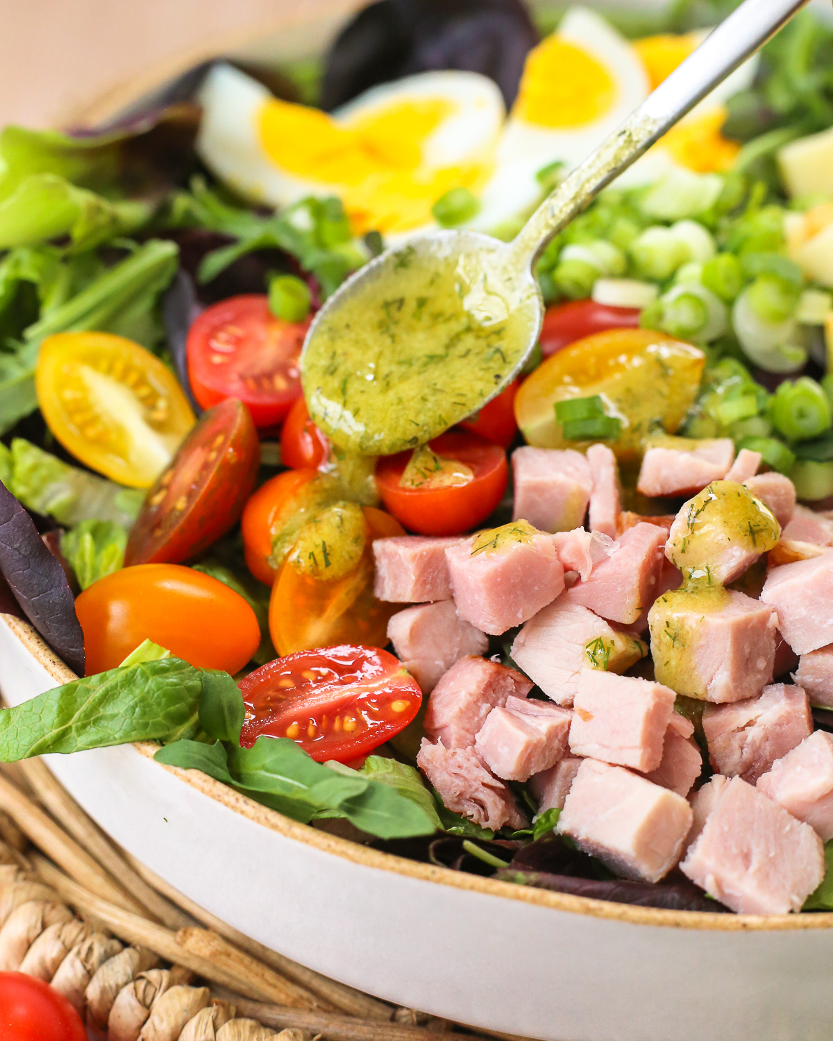 A small silver spoon drizzles a ranch vinaigrette over the top of a chef's salad made with leftover ham and colorful fresh vegetables.