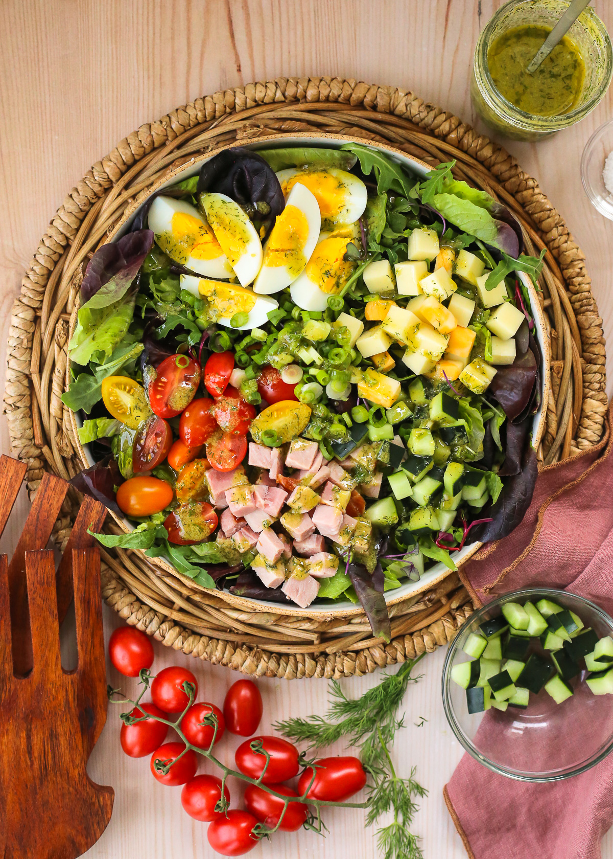 A colorful chef's salad with ham, tomatoes, cucumbers, hard boiled eggs, cheese, and a ranch vinaigrette dressing, served in a ceramic salad bowl on a woven straw placemat.