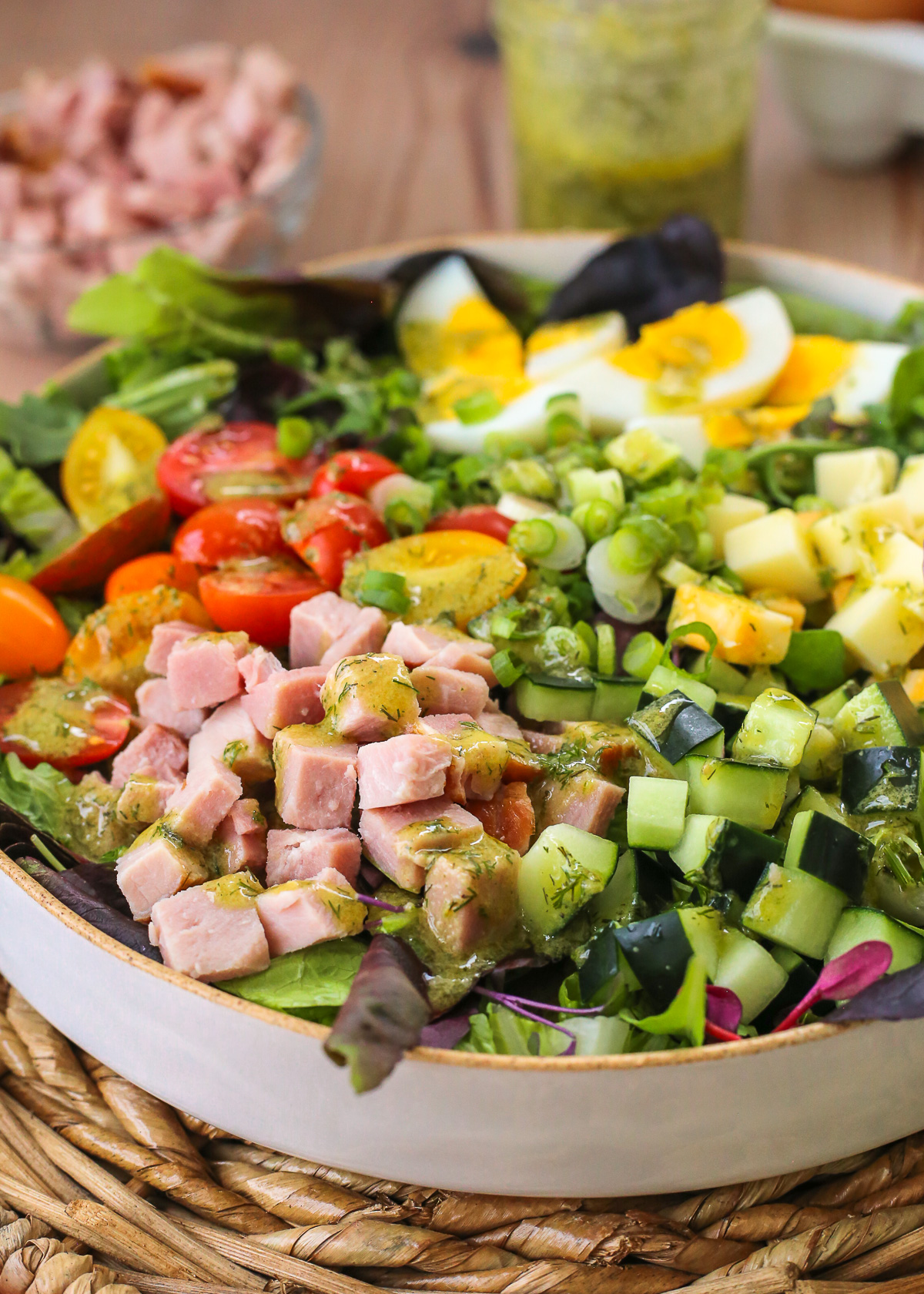 A colorful chef's salad with ham, cucumbers, tomatoes, cheese, hard boiled eggs, and sliced green onions displayed in a large ceramic salad bowl on a wooden kitchen table.