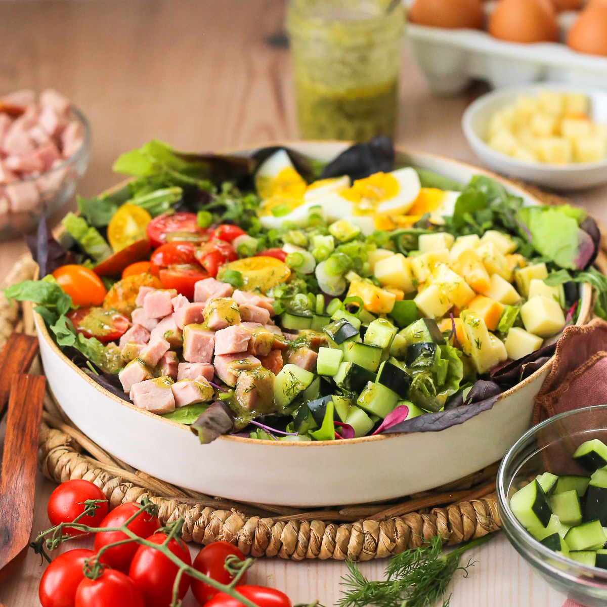 A chef's salad with ham and a variety of colorful fresh vegetables, served in a ceramic salad bowl with extra dressing and salad serving tongs in the background.