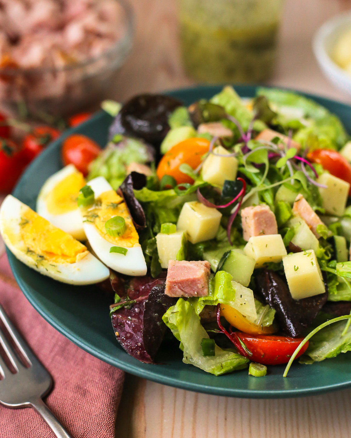 A chef's salad with ham and ranch vinaigrette dressing served on a dark green ceramic salad bowl, with a fork placed nearby.