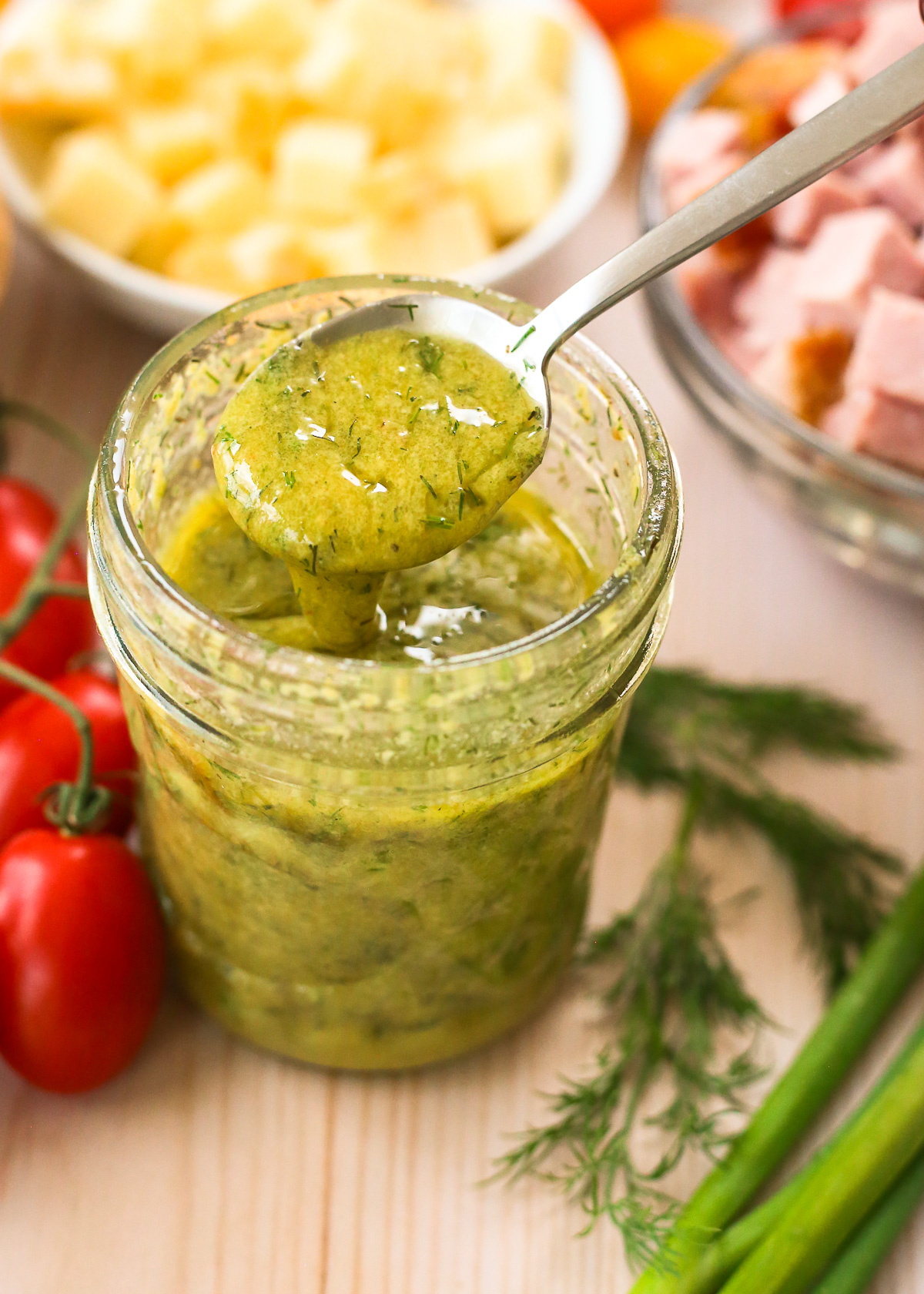 A small silver spoon lifts a scoop of homemade salad dressing from a small glass jar, with specks of fresh dill visible in the dressing mixture.
