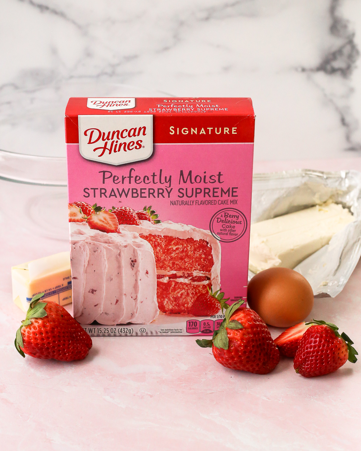 A box of Duncan Hines brand strawberry cake mixed, displayed on a kitchen countertop with fresh strawberries, one egg, one stick of butter, one block of cream cheese, and a clear glass mixing bowl.