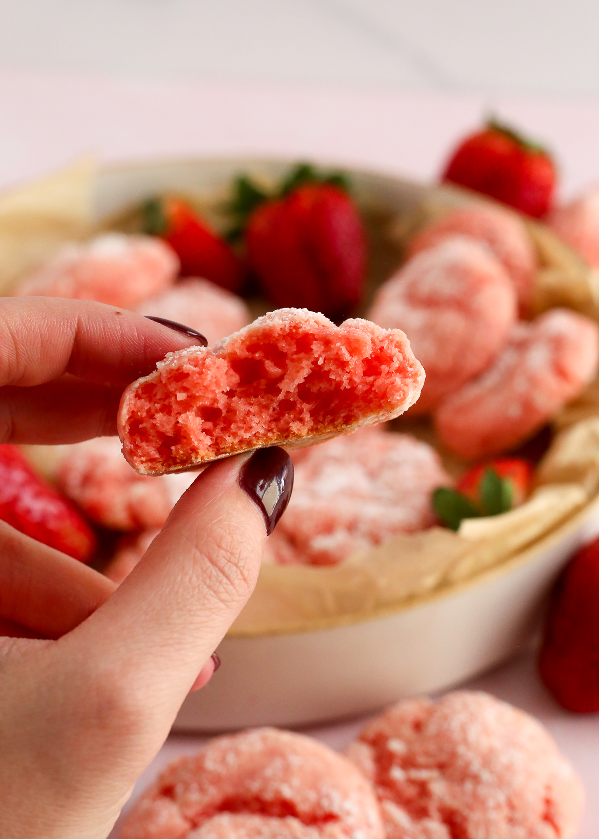 A woman's hand holds a strawberry gooey butter cookie, broken in half, to reveal the fluffy pink interior.