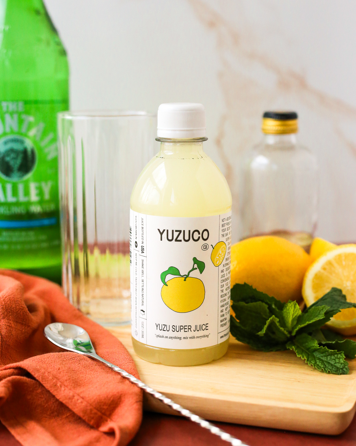 A bottle of yuzu juice displayed on a brightly lit kitchen countertop along with lemons, fresh mint, an empty glass, and a bottle of sparkling water.