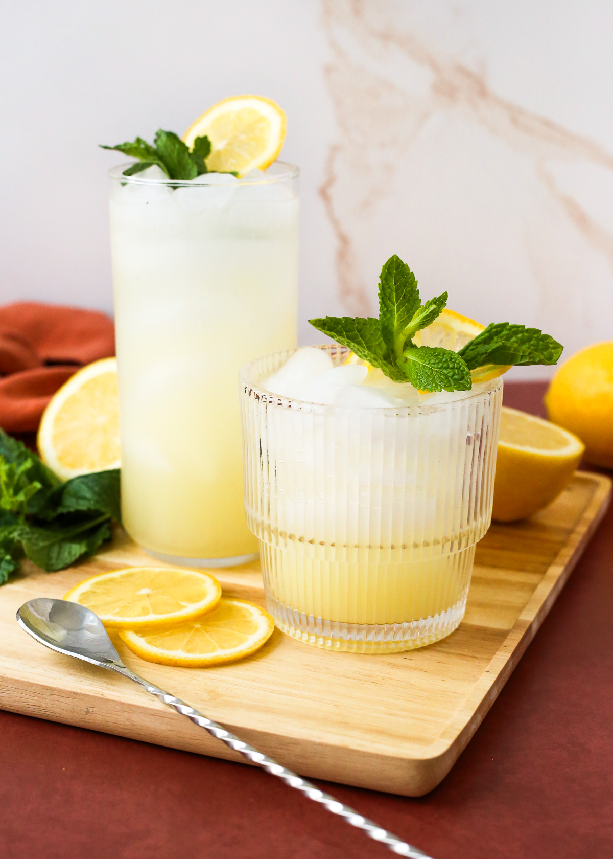 Two glasses of sparkling yuzu lemonade garnished with fresh mint and lemon slices served on a wooden tray in a brightly lit kitchen.