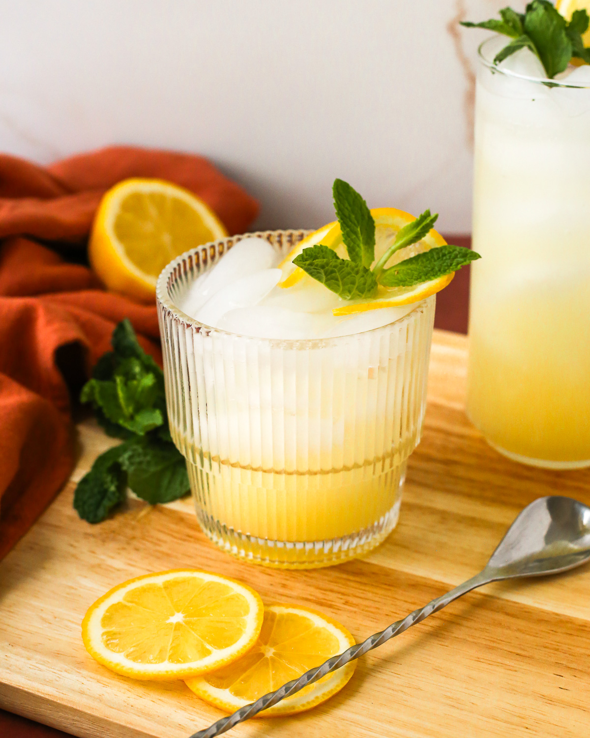 A glass of sparkling yuzu lemonade garnished with fresh mint and a lemon slice served on a wooden tray, with sliced lemons in the background.