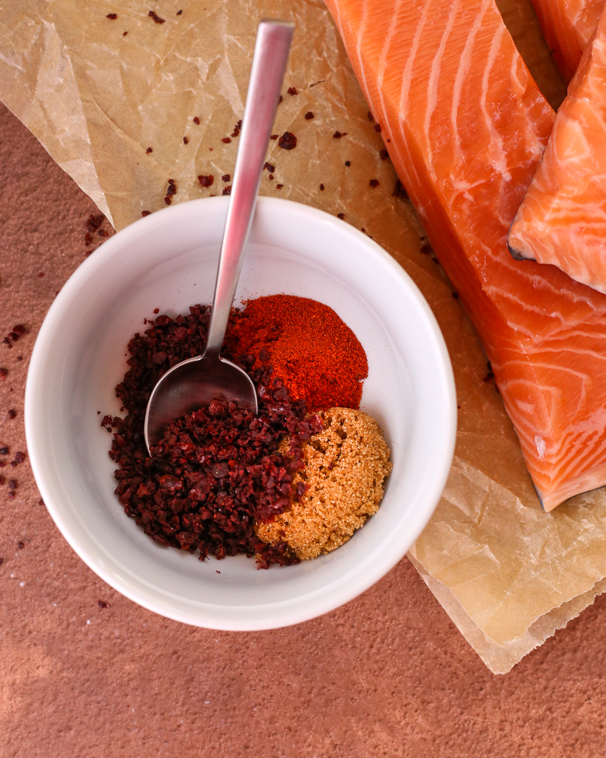 A small white prep bowl containing ground sumac, smoked paprika, and brown sugar with a small spoon on a kitchen countertop.