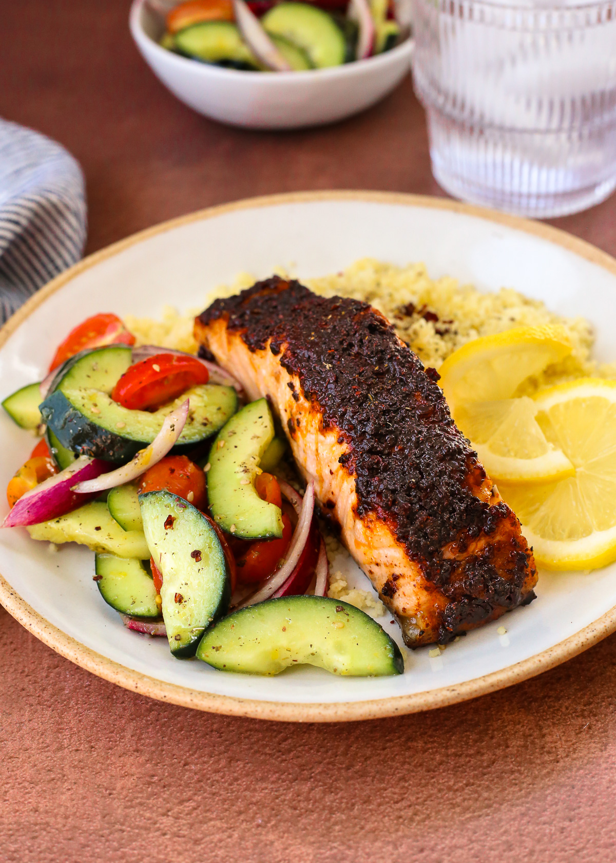 A dinner plate with a serving of sweet sumac salmon served over couscous and a cucumber, tomato, and red onion side salad, garnished with lemon slices.