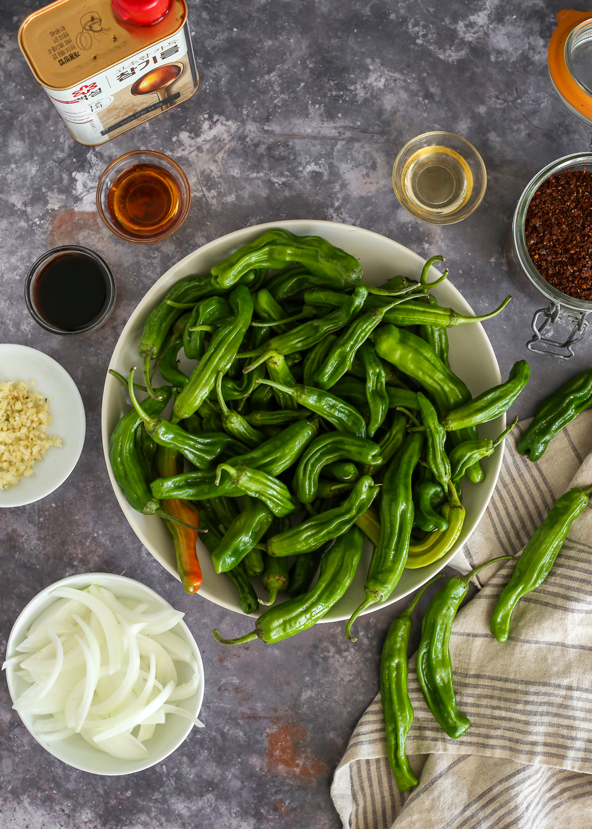 Fresh shishito peppers displayed on a kitchen counterstop along with sliced onion, minced garlic, soy sauce, sesame oil, and red pepper flakes.