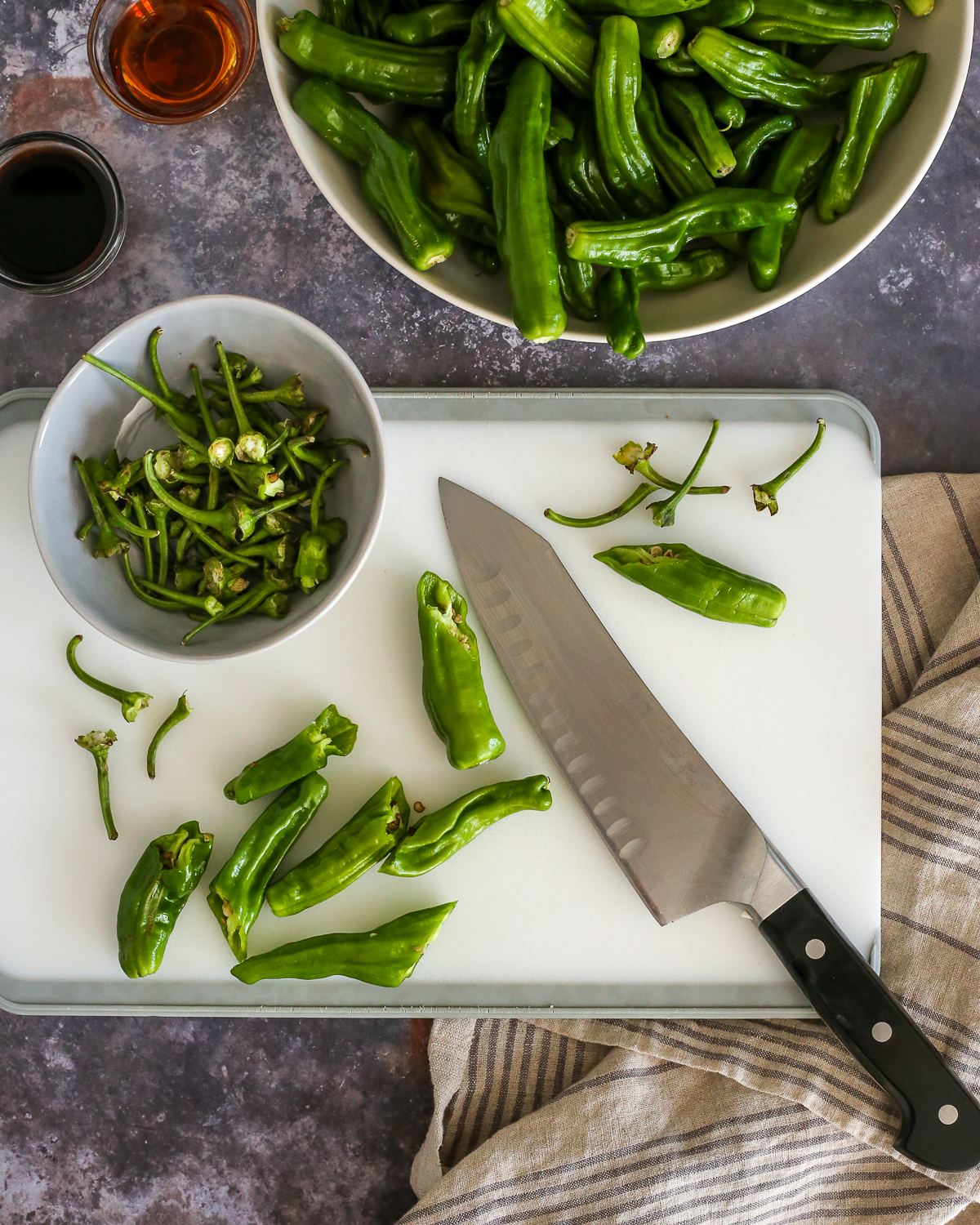 A white cutting board with shishito peppers scattered across it, with stems removed and discarded in a small ceramic prep bowl placed nearby.