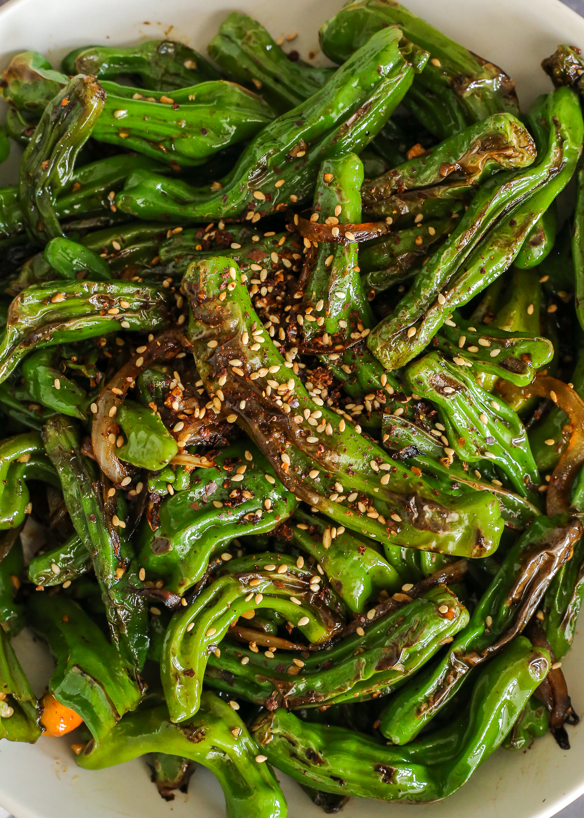 A close view of the finished version of blistered shishito peppers, with dark char marks, a light sauce, and sesame seeds and red pepper flakes on top as garnish.