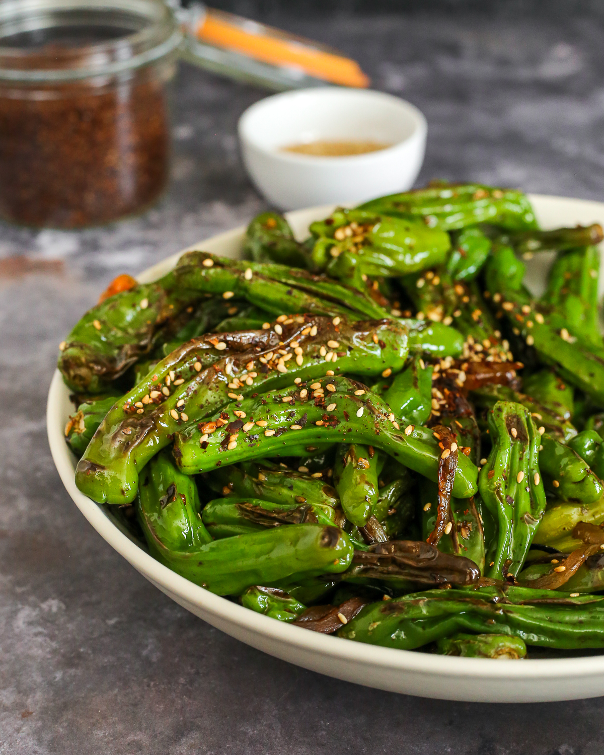 A large serving bowl full of blistered shishito peppers garnished with sesame seeds and red pepper flakes.