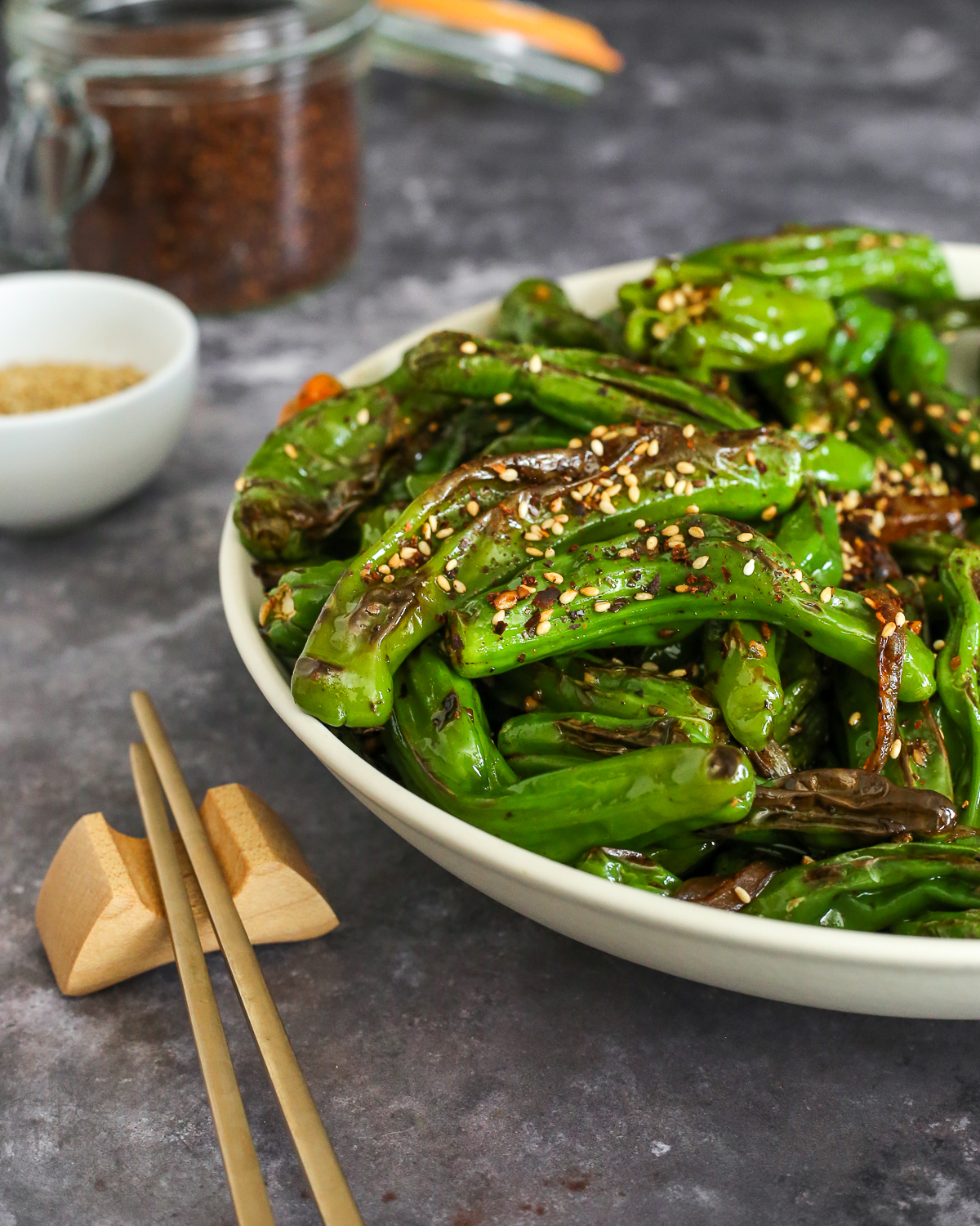 A large bowl of blistered shishito peppers served on a kitchen countertop with a pair of chopsticks placed nearby and a small white ramekin filled with sesame seeds in the background.