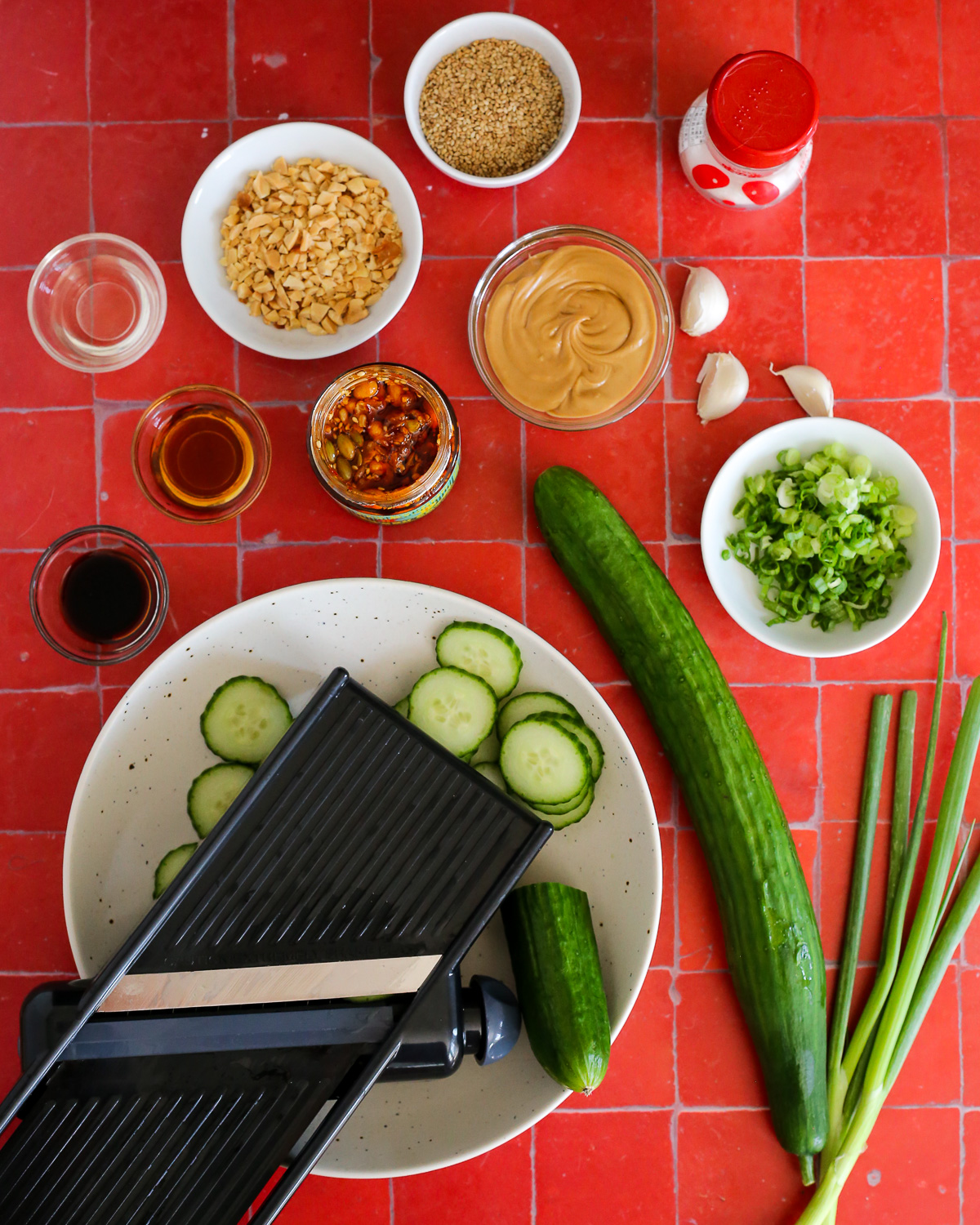 An assortment of ingredients needed to make a cucumber salad with peanut butter and chili crisp, including cloves of garlic, sliced green onions, various seasonings and spices, sesame oil, and a fresh cucumber partially sliced with a mandoline.
