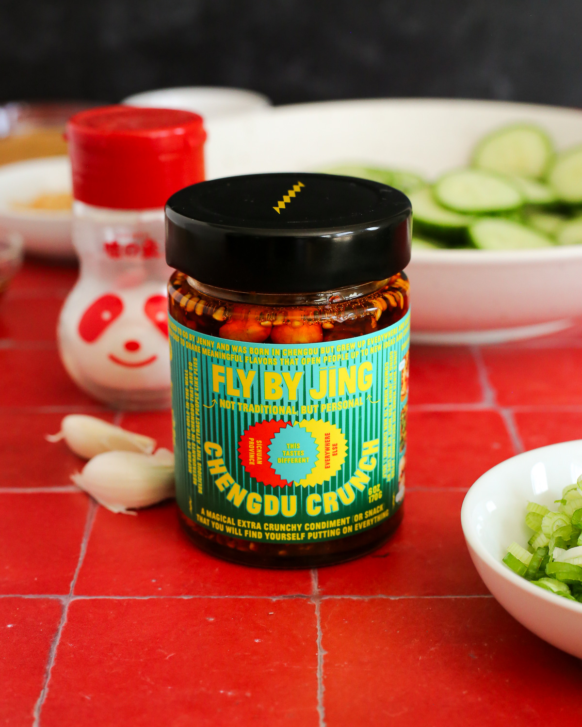 A jar of chili crisp is displayed on a kitchen countertop, with the label reading "Fly By Jing Chengdu Crunch" in block letters, yellow on a green background.
