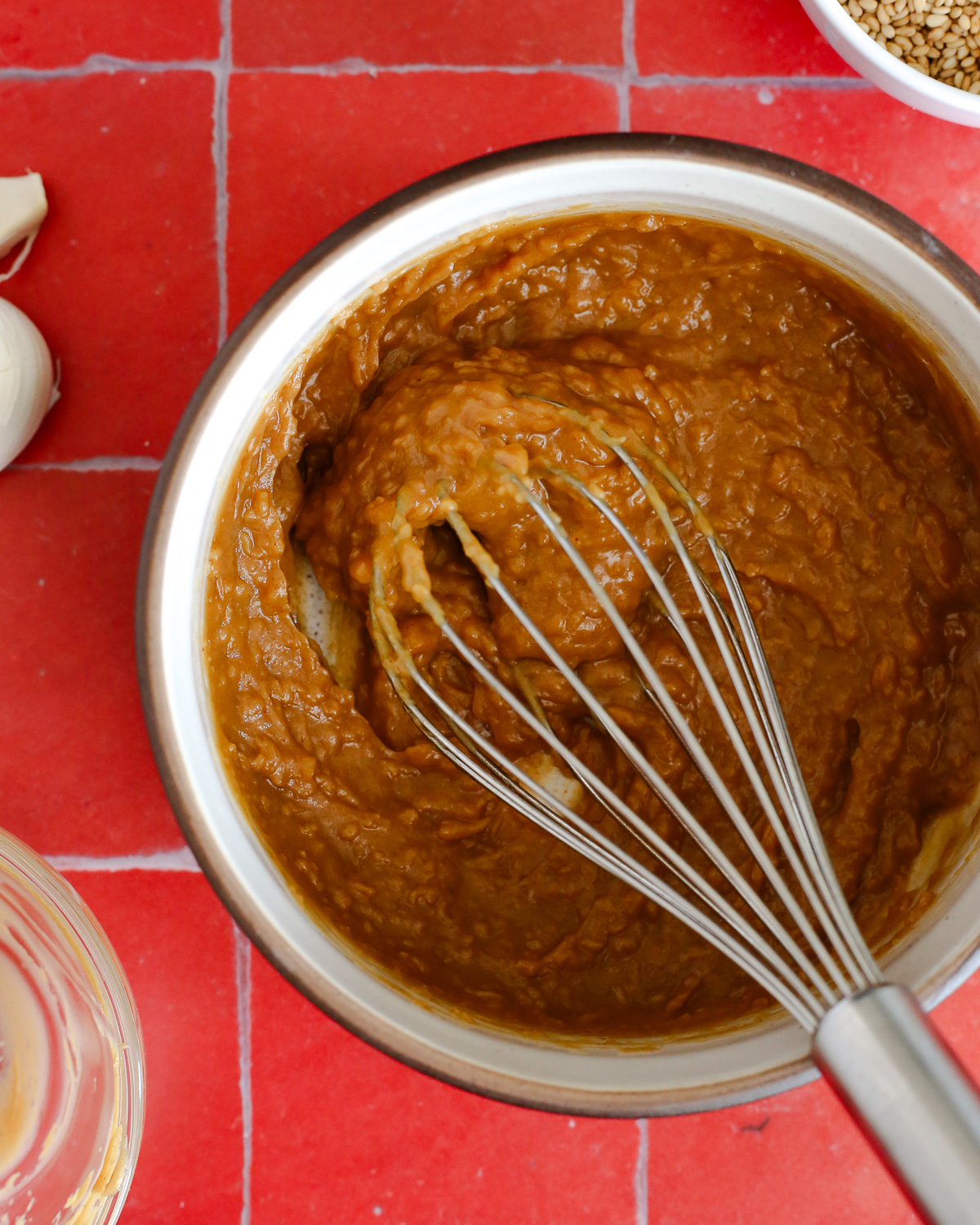 A small mixing bowl contains a thick, creamy peanut sauce with a small metal whisk resting on the side.