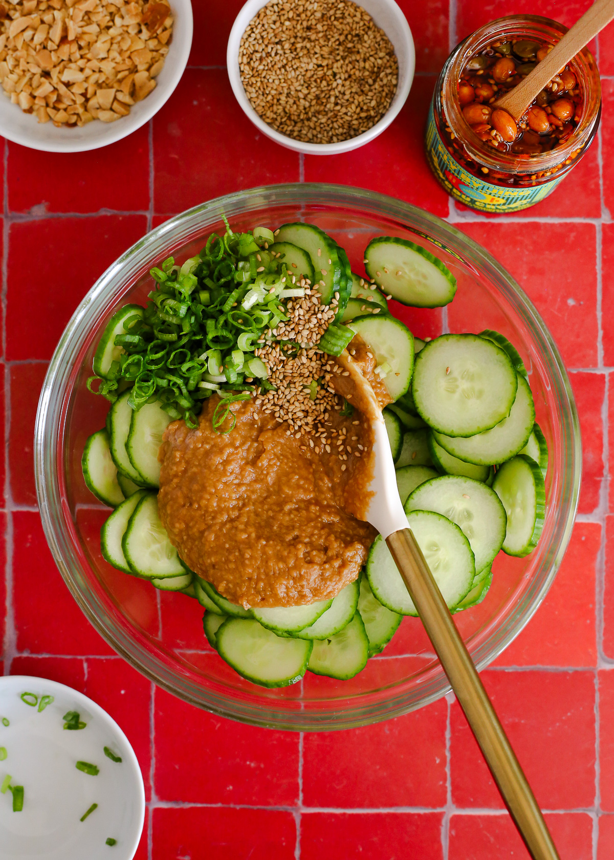 An overhead view of a large mixing bowl filled with sliced cucumbers, sliced green onions, and a freshly mixed homemade peanut sauce.