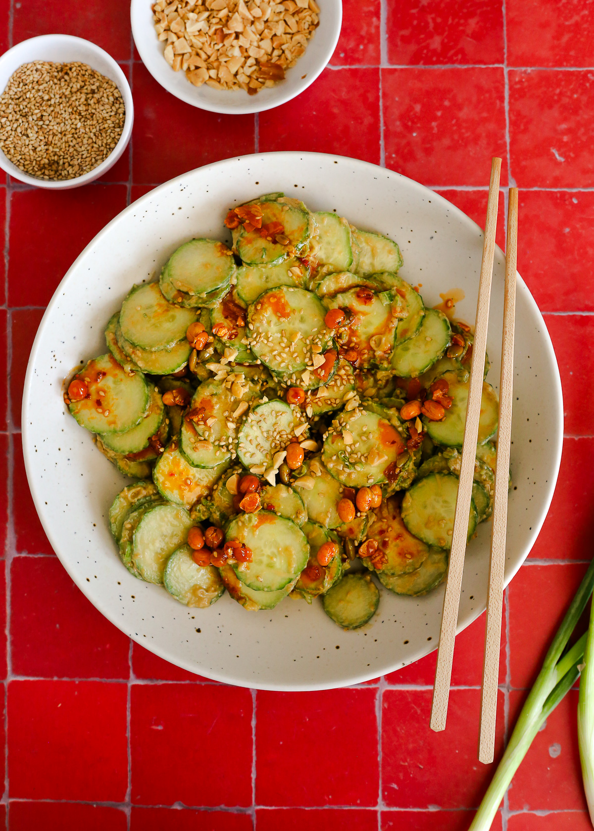 Overhead view of a cucumber salad recipe made with a peanut dressing, drizzled with chili crisp and topped with sliced green onions, chopped peanuts, and sesame seeds.
