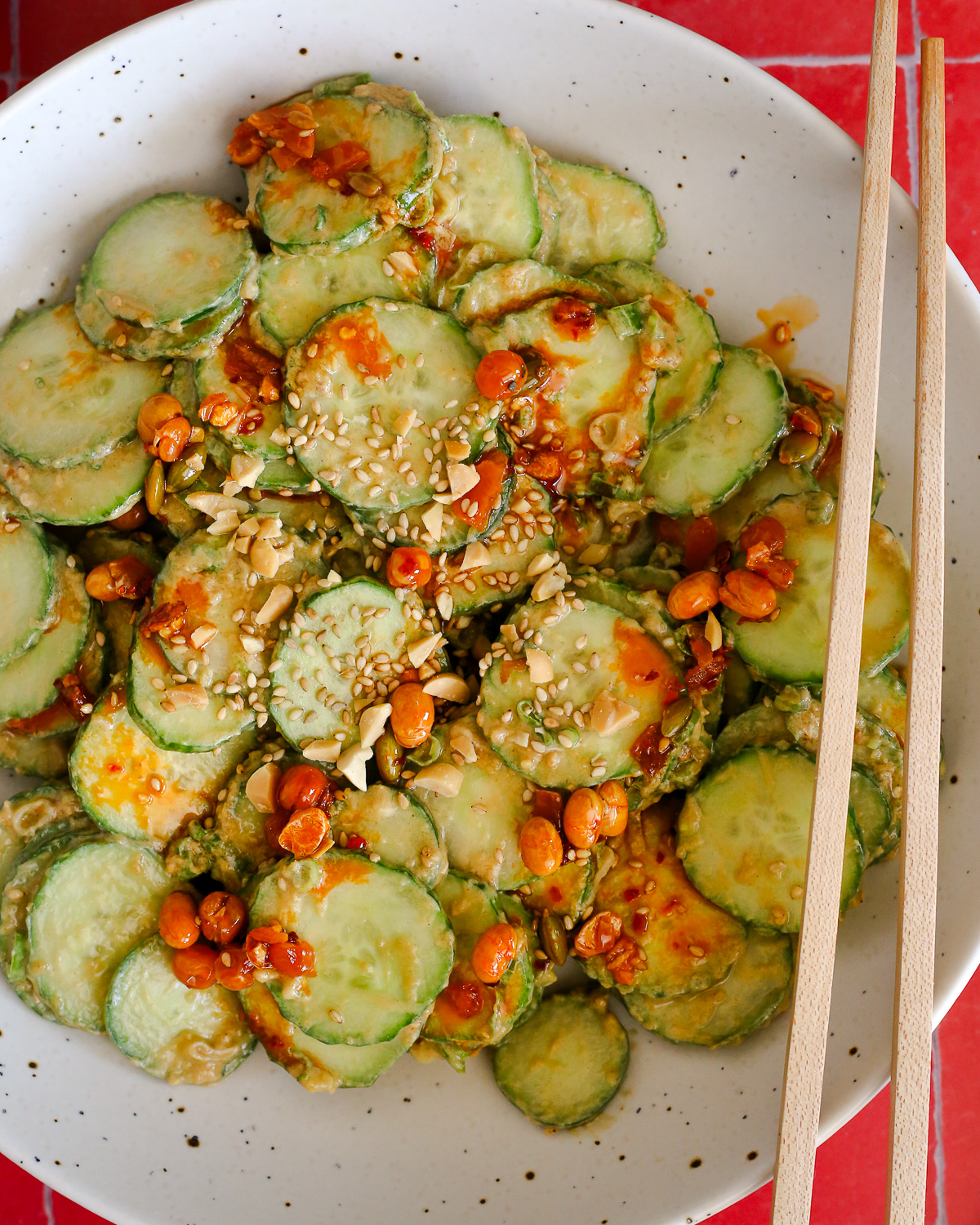 A close view of a cucumber salad with peanut dressing and chili crisp, topped with chopped peanuts, sliced green onions, and sesame seeds with a pair of wooden chopsticks resting on the side of the bowl.