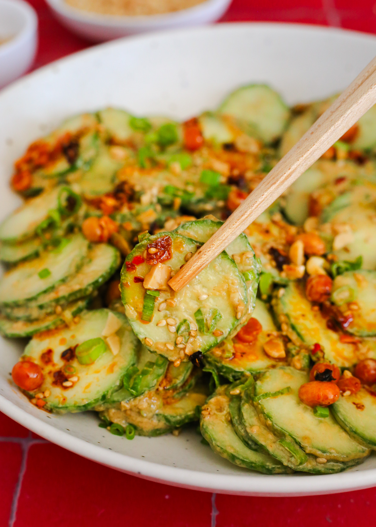 A pair of wooden chopsticks lifts a bite of sliced cucumber salad from a serving bowl, coated in a creamy peanut butter dressing with chili crisp, green onions, and sesame seeds.