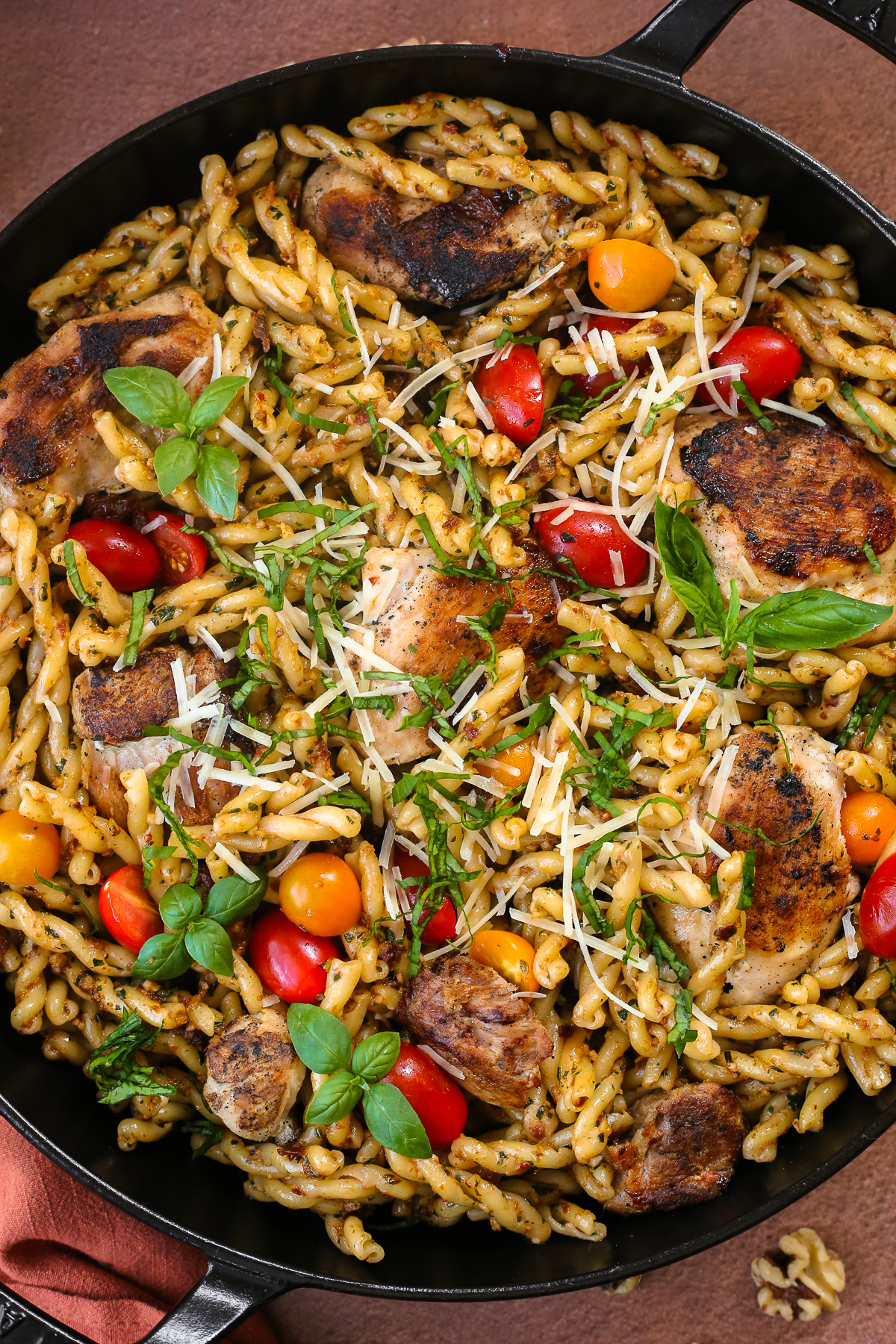 A close view of a pesto rosso pasta skillet, with seared chicken thighs and cherry tomatoes mixed throughout, topped with fresh basil and parmesan cheese.
