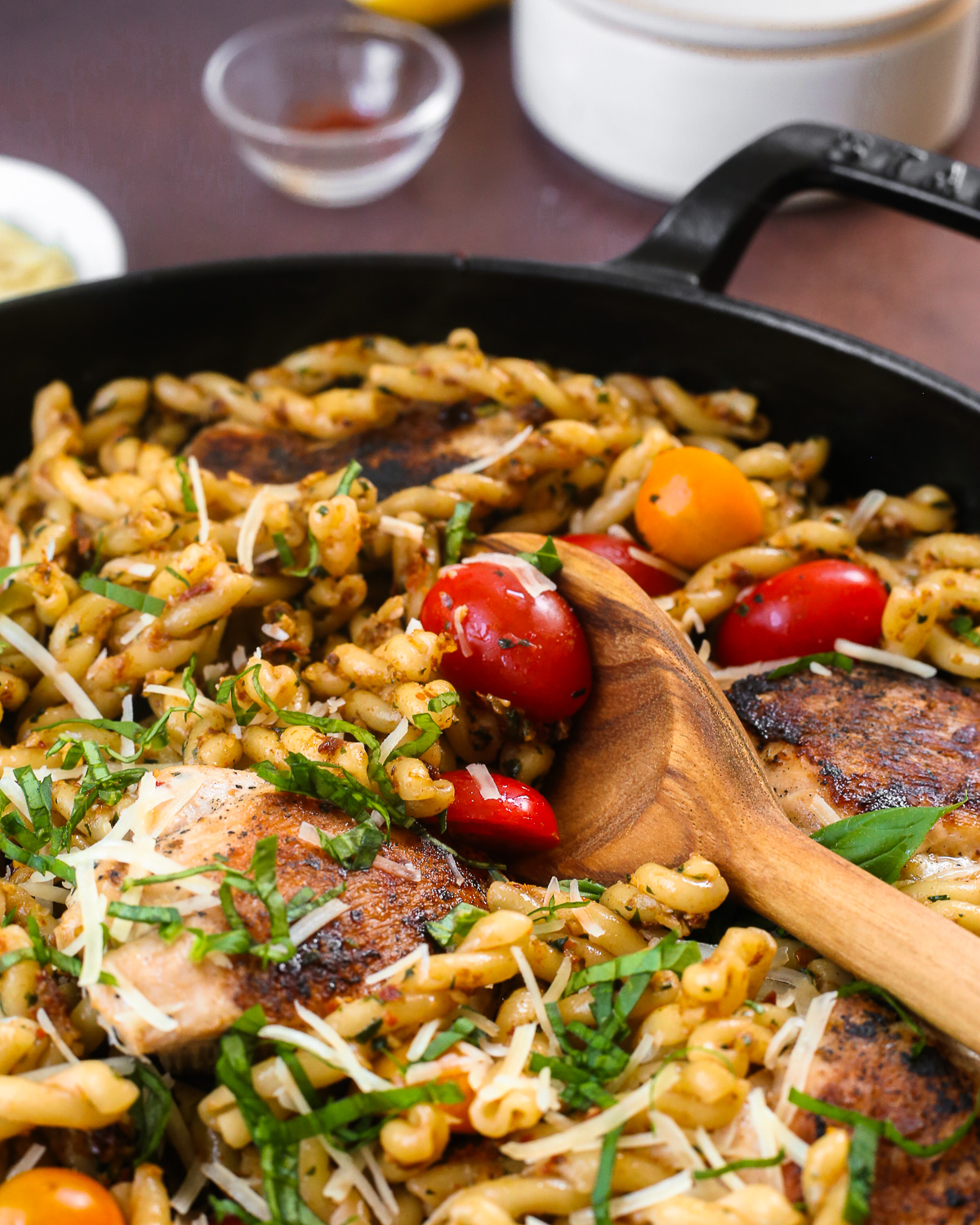 A wooden serving spoon scoops a serving of sun-dried tomato pasta from a skillet that also contains seared chicken and cherry tomatoes.