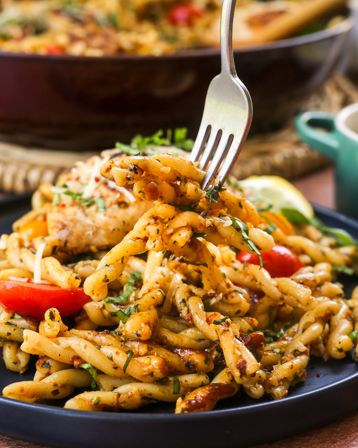 A silver fork lifts a large bite of pesto rosso pasta from a dinner plate that also contains a piece of seared chicken and cherry tomato halves.
