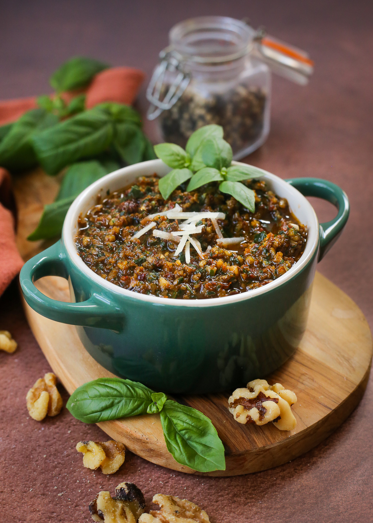 A green jar of pesto rosso, or sun-dried tomato pesto, displayed on a small wooden serving board, garnished with grated permensan cheese and fresh basil leaves. 