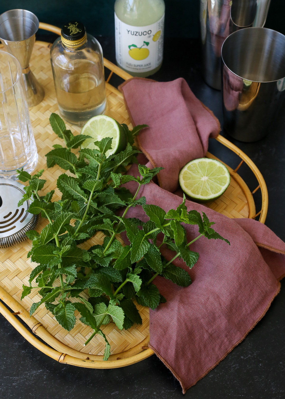 A large handful of fresh mint displayed on a serving tray along with a lime, empty cockail glasses, a bottle of yuzu juice, a bottle of simple syrup, and a Boston shaker. 