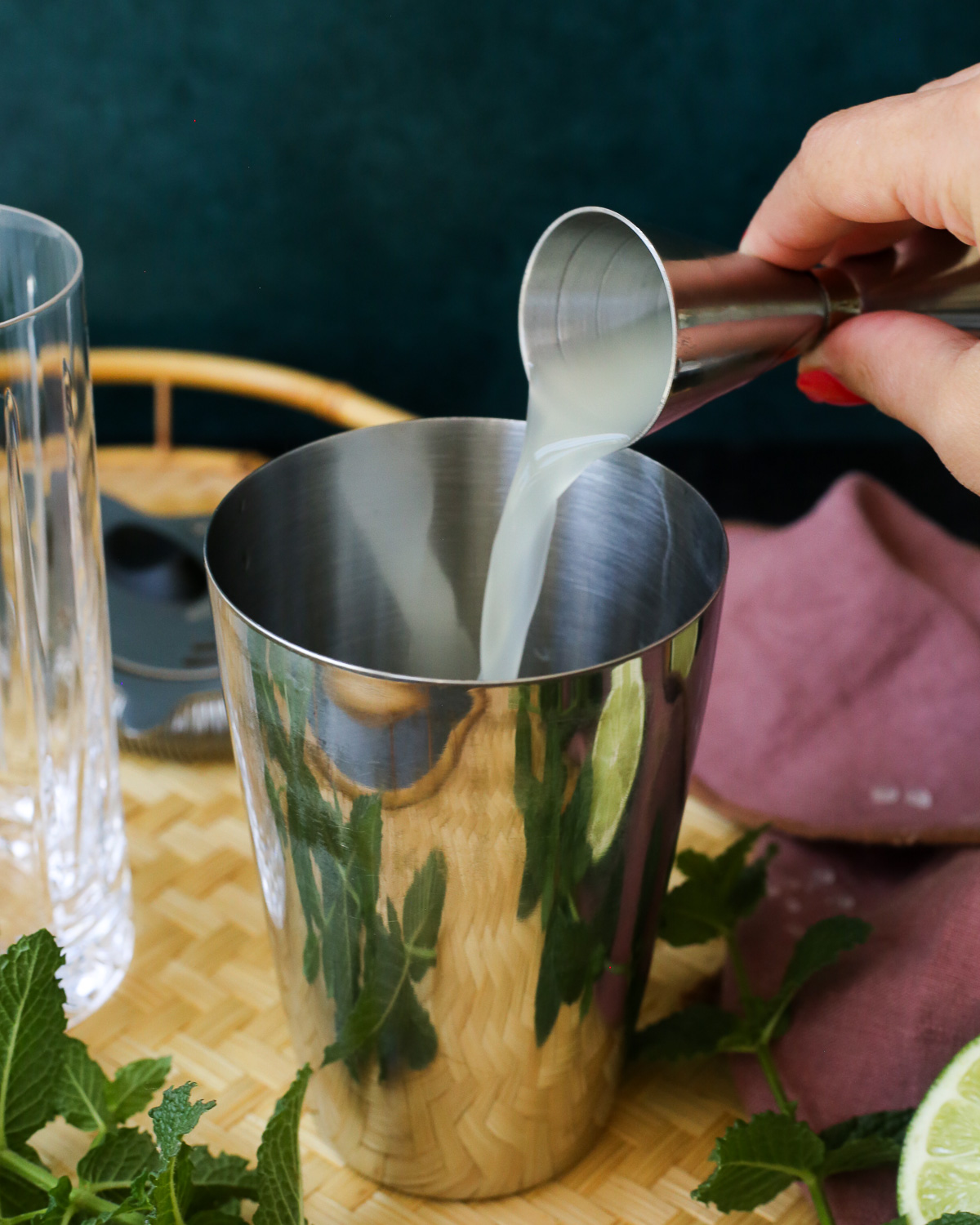 A woman's hand pours yuzu juice into a cocktail shaker using a silver jigger.