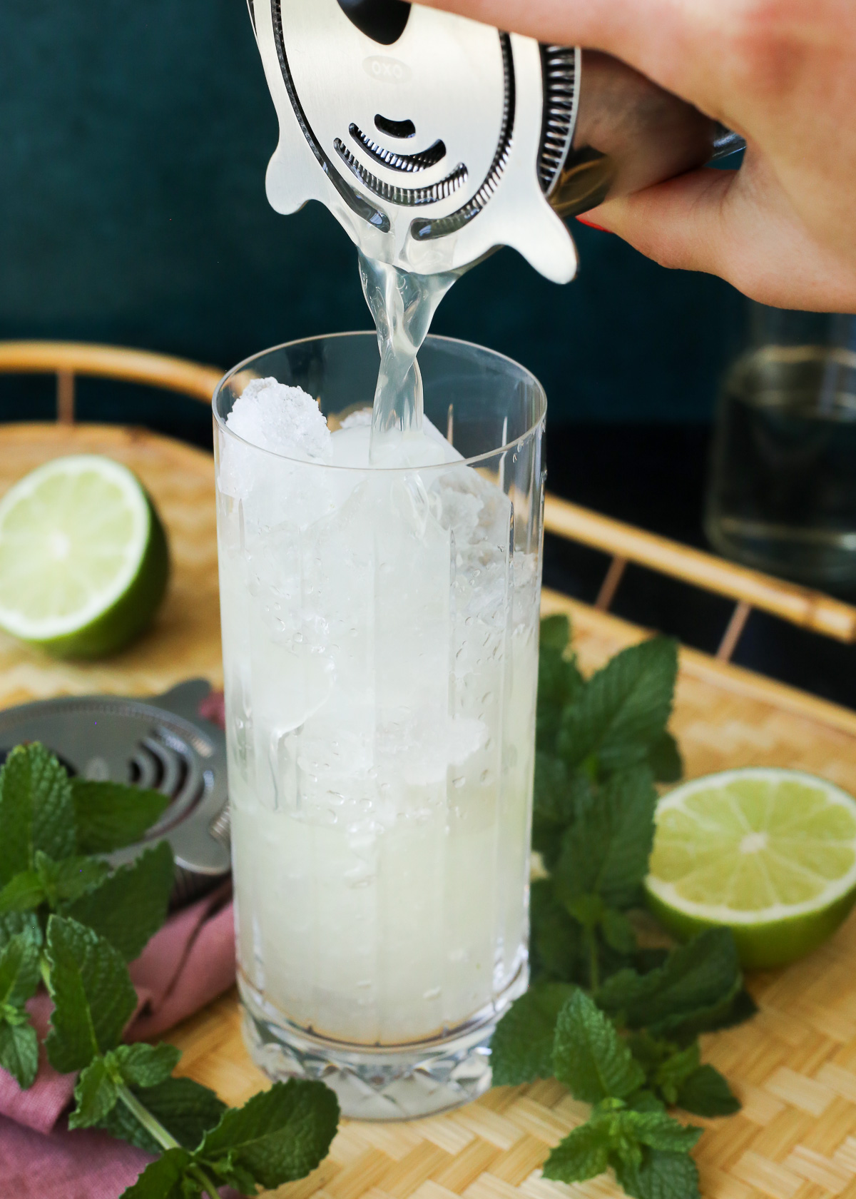 A woman's hand strains a yuzu mojito mix into a highball glass filled with crushed ice.