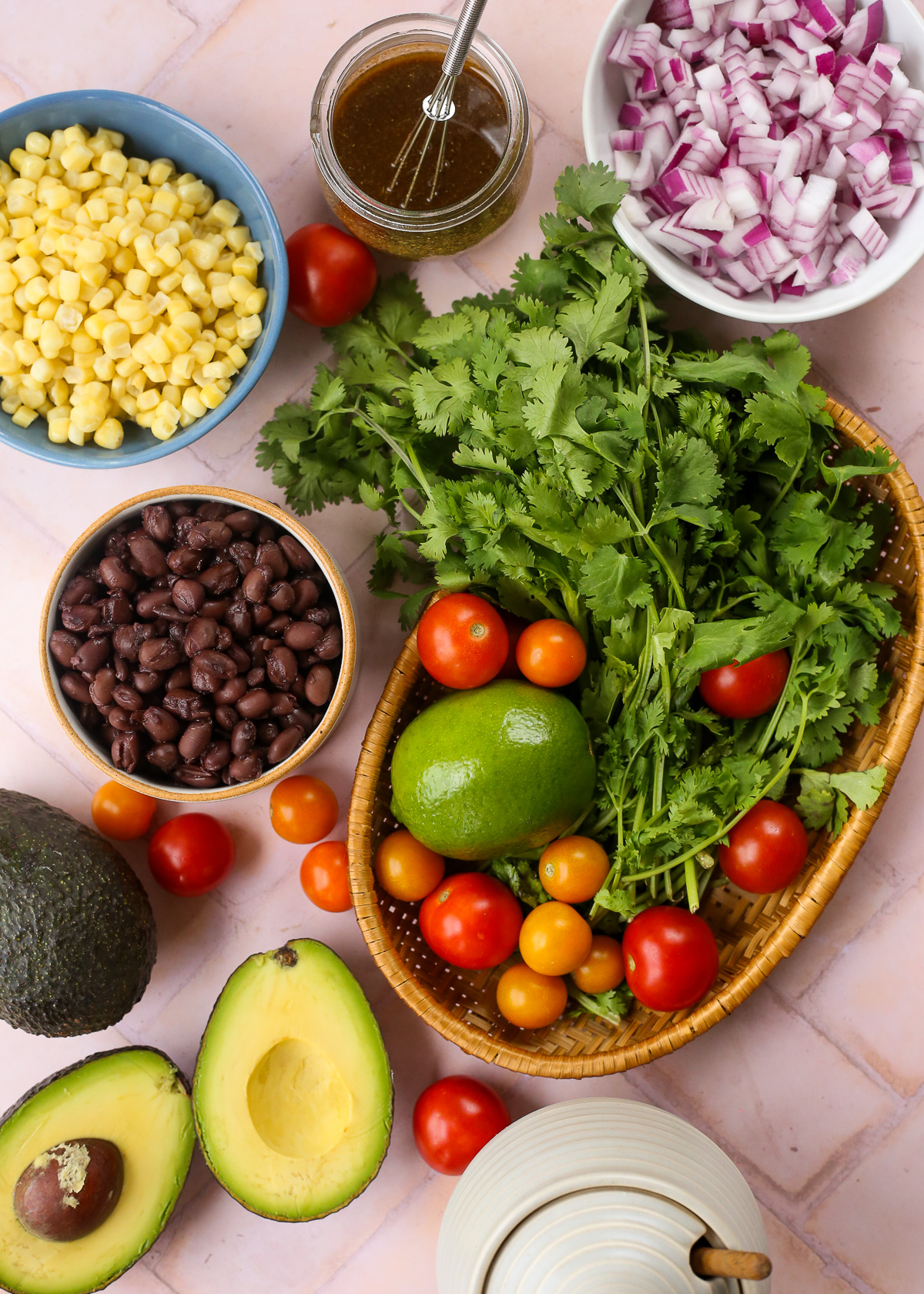 An assortment of ingredients spread on a stone countertop, including corn, canned black beans, diced red onion, fresh cilantro, cherry tomatoes, a lime, and avocados. 
