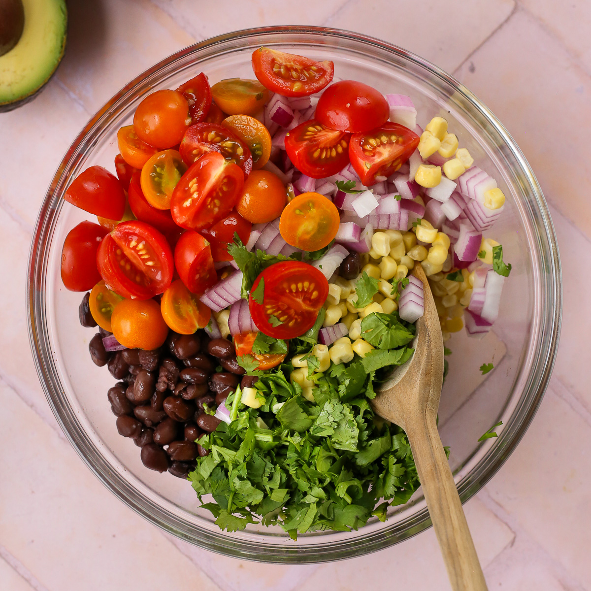 An unmixed version of an avocado black bean salad in a large glass mixing bowl, with a wooden spoon resting on one side. 