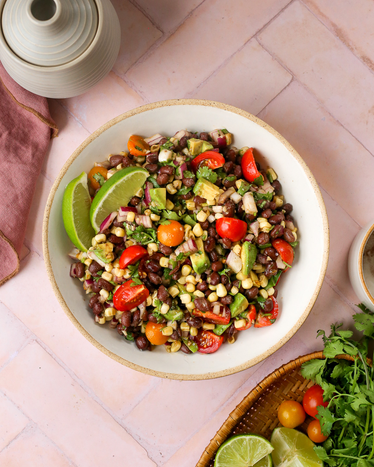An overhead view of an avocado black bean salad, mixed together with corn, tomatoes, red onion, and cilantro, served in a large ceramic bowl with lime wedges. 