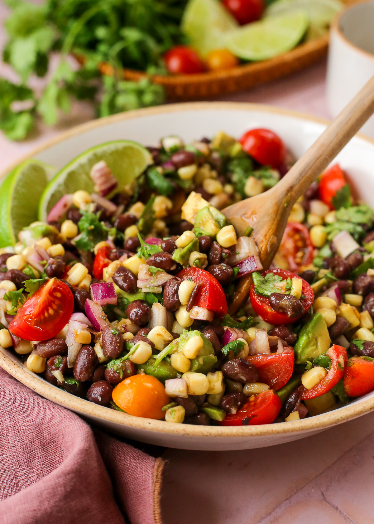 A wooden spoon lifts a scoop of a dense bean salad made with black beans, corn, tomatoes, and avocado from a ceramic serving bowl. 