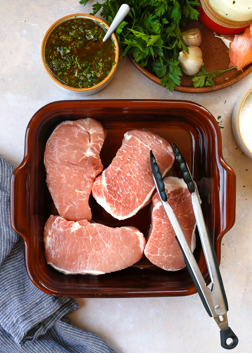 Four boneless pork chops in a shallow baking dish, with a dish of chimichurri marinade nearby and a pair of grilling tongs resting on the side of the dish. 