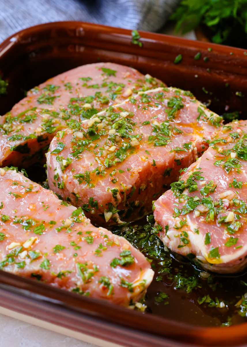 Four uncooked pork chops marinate in a chimichurri marinade. 