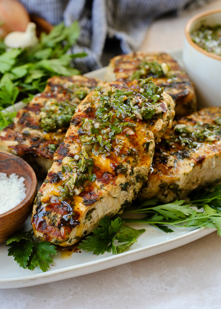 Chimichurri pork chops served on a large platter, with prominent grill marks and a small dish with extra chimichurri in the background. 