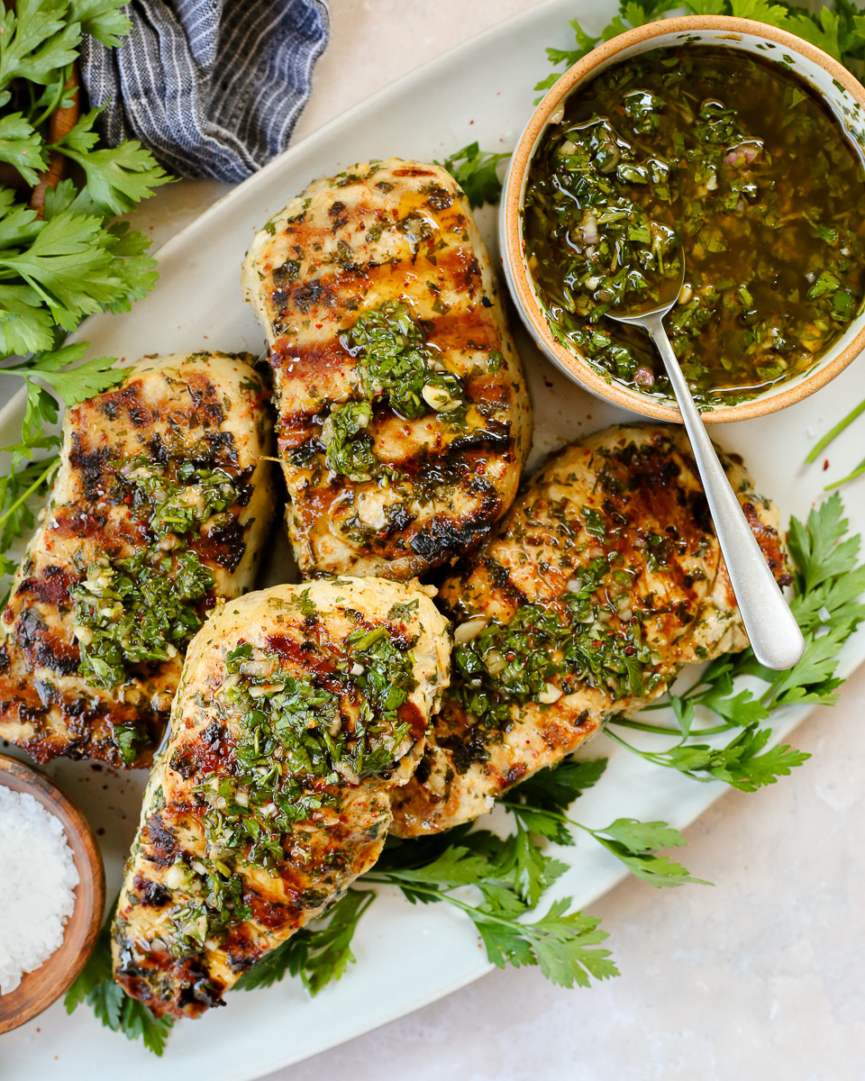 Four grilled pork chops topped with chimichurri, served on a platter with a small dish holding extra chimichurri sauce. 