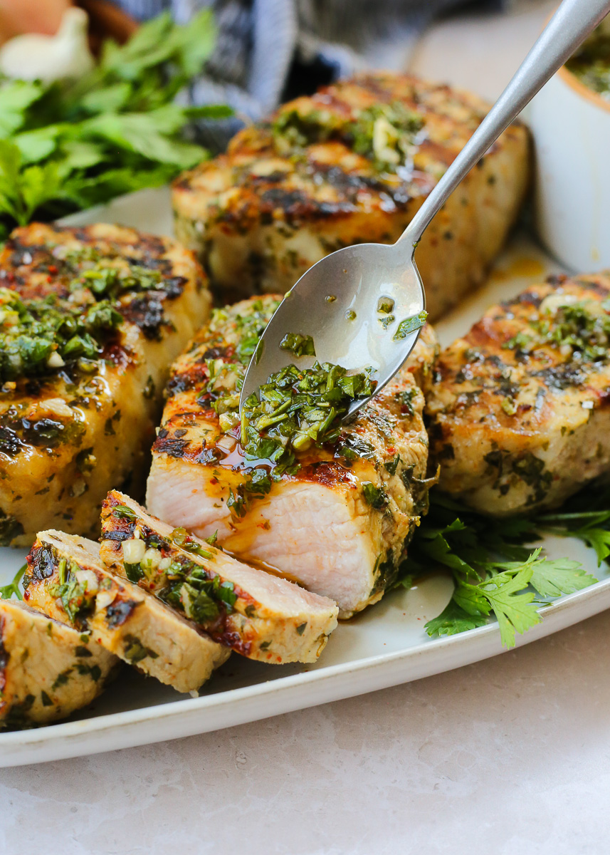 A spoon drizzles chimichurri sauce over grilled pork chops, which are sliced open to show the juicy, tender interior. 