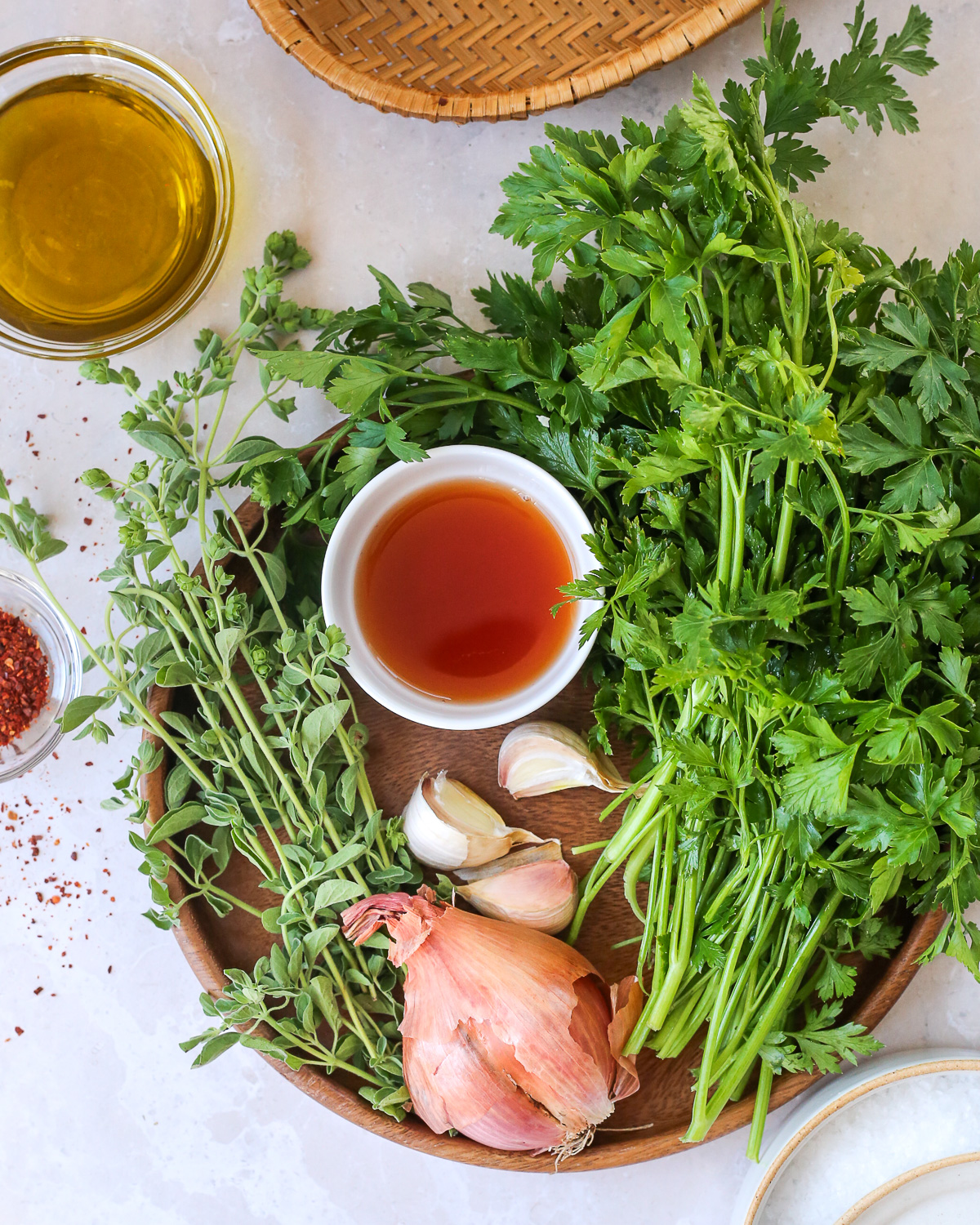 A large bunch of fresh parsley on a wooden plate, along with fresh oregano, a few garlic cloves, a shallow, and a small white prep bowl filled with red wine vinegar. 
