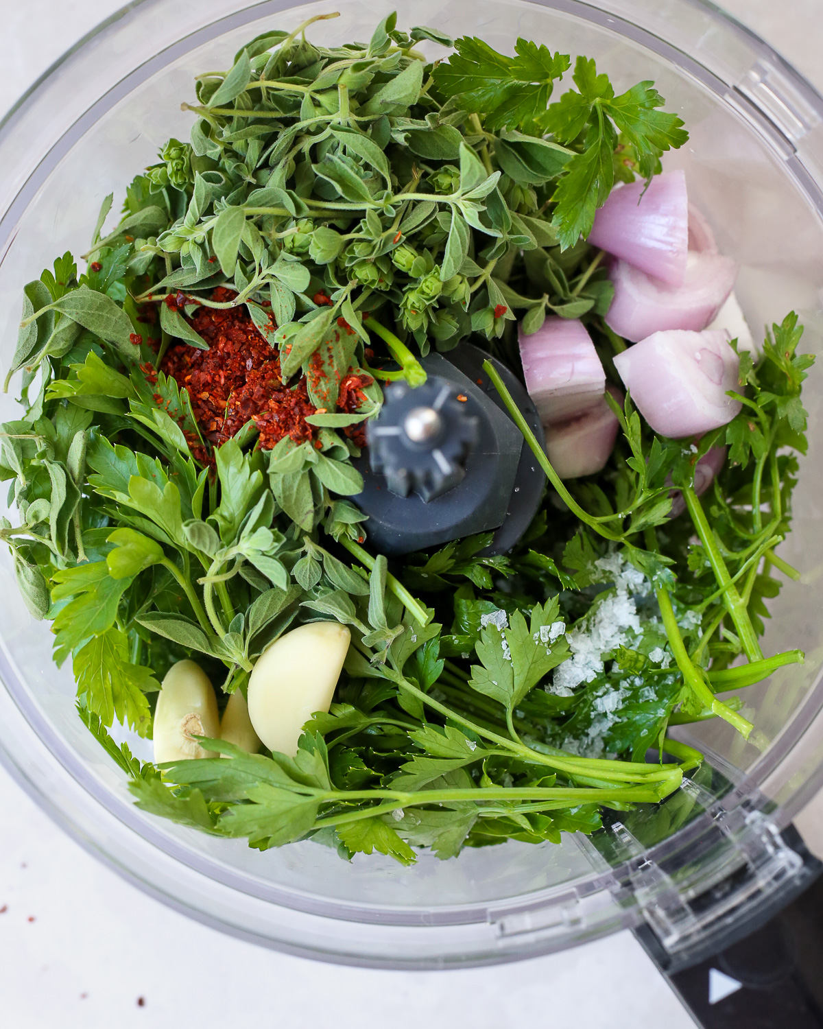 A view into the bowl of a food processor, showing fresh parsley, fresh oregano, a few cloves of garlic, and a chopped shallot along with a pinch of salt and red pepper flakes. 
