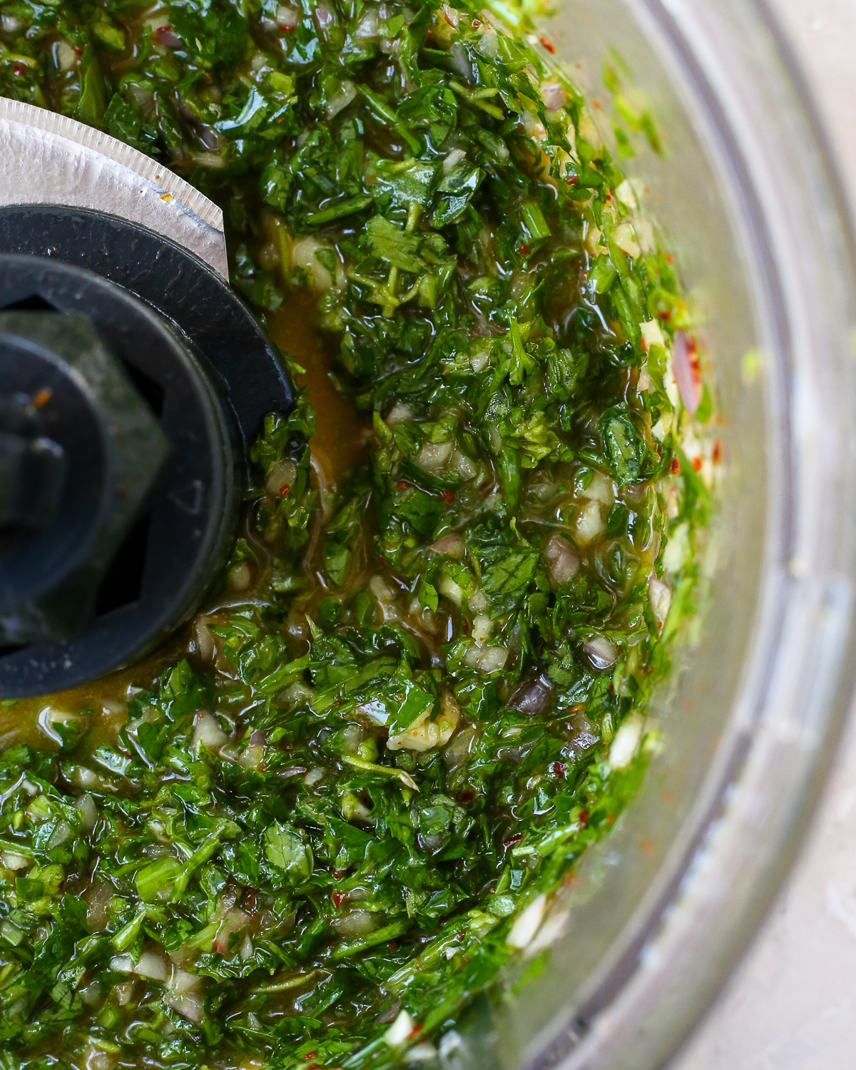 A closeup of the blades of a food processor, showing the loose texture and chunky consistency of chimichurri sauce. 