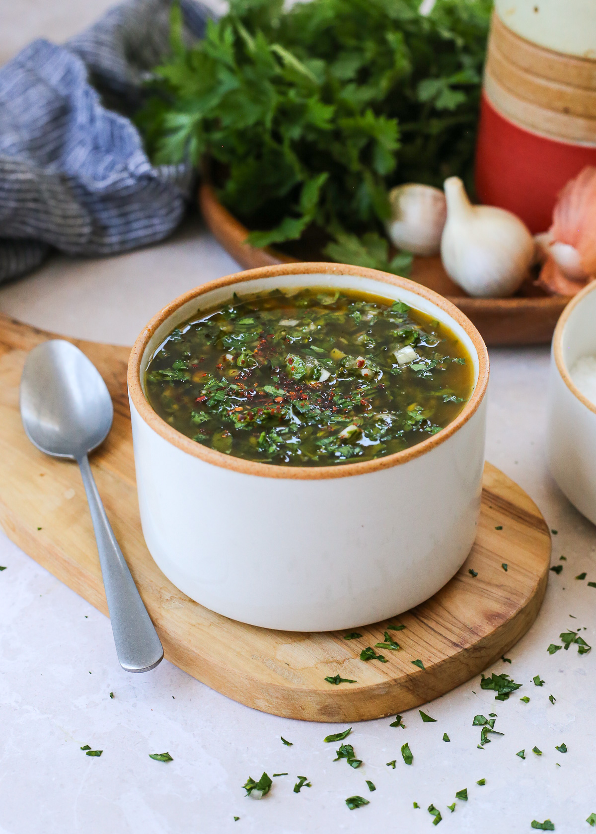 A small serving bowl full of chimichurri sauce rests on a small wooden serving tray on a kitchen countertop with extra ingredients scattered in the background. 