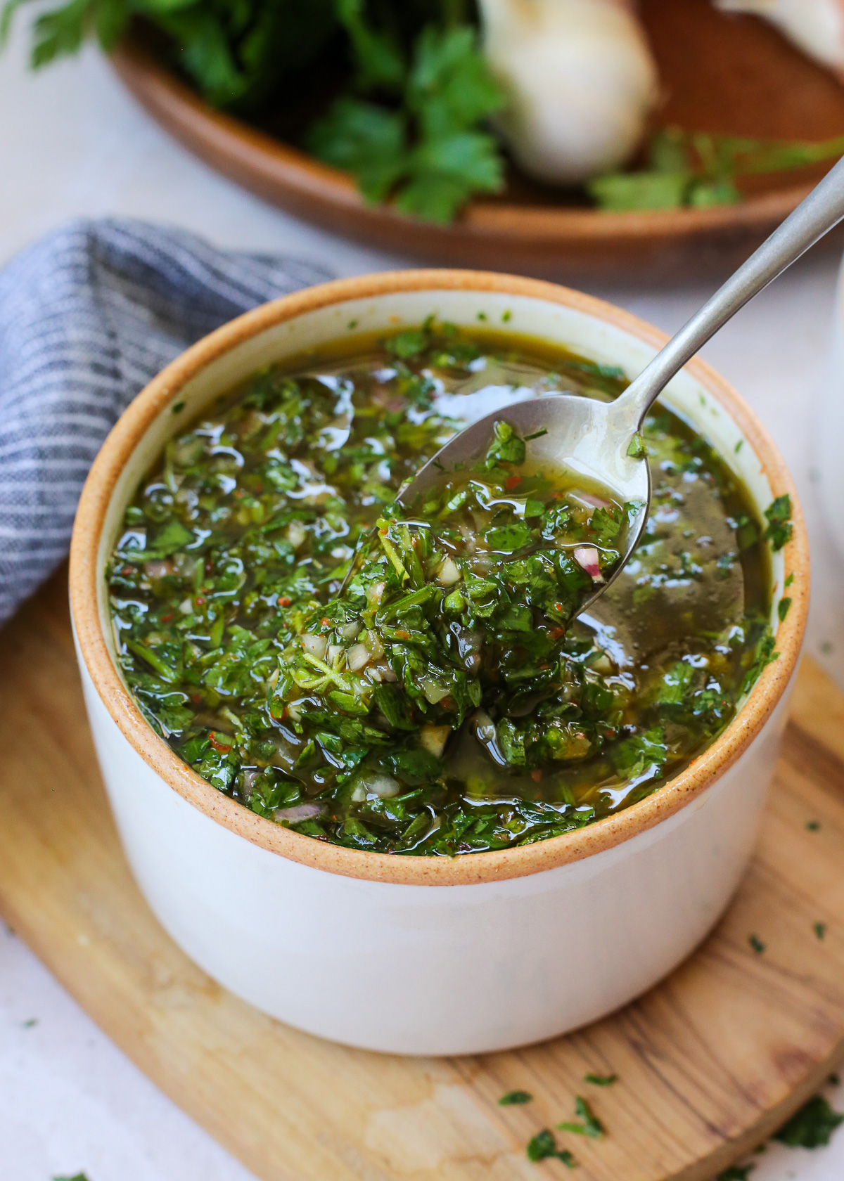A spoon lifts a scoop of chimichurri sauce from a ceramic bowl, showing the green herbs, minced garlic, and red pepper flakes. 