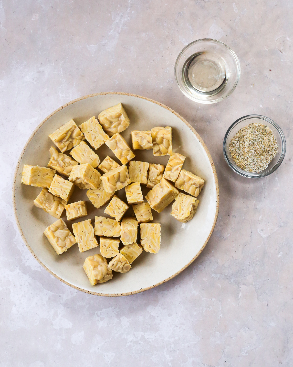 Uncooked tempeh, cut into bite sized cubes, displayed on a plate along with prep bowls containing cooking oil and an all-purpose seasoning blend.