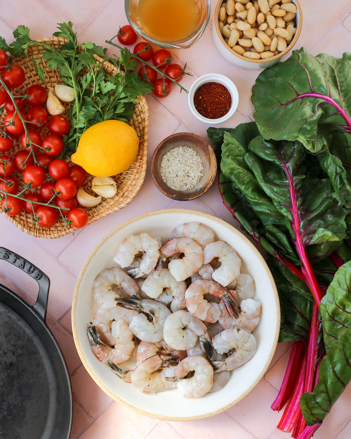An assortment of ingredients to make a shrimp skillet recipe, including raw peeled shrimp, cherry tomatoes, large leaves of Swiss chard, a lemon, parsley, garlic cloves, white beans, seasonings, and broth. 