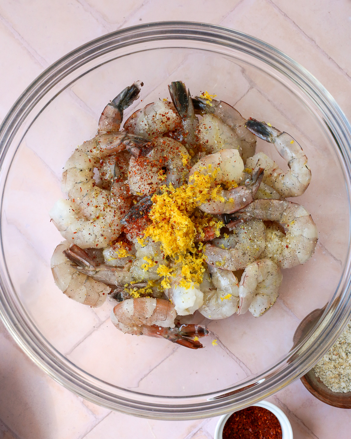 A glass mixing bowl filled with raw shrimp, with seasonings added on top. 