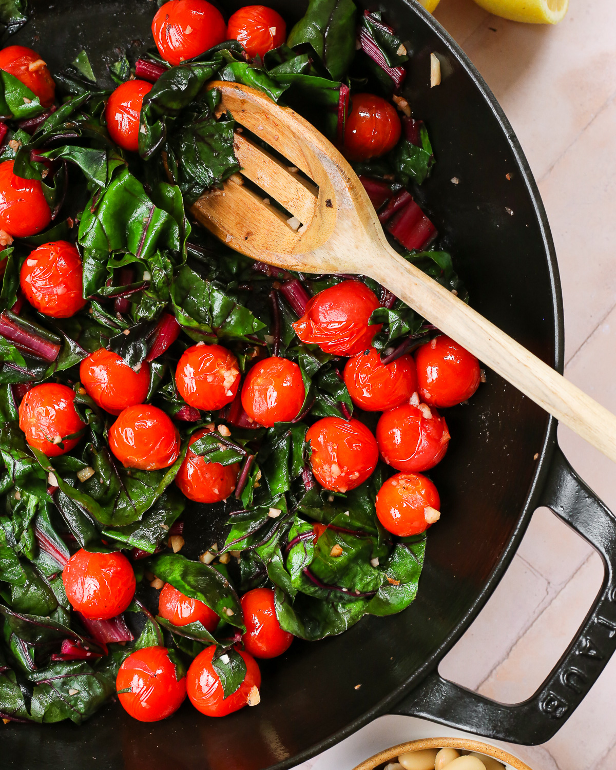 A wooden spoon stirs a mixture of blistered tomatoes and cooked greens, braising with a garlicky broth. 